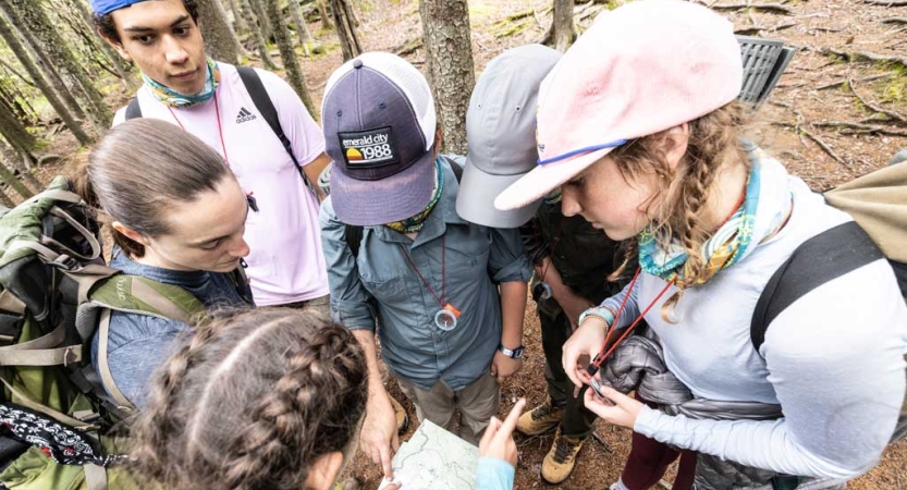 A group of young people wearing backpacks stand in a circle and examine a map. 